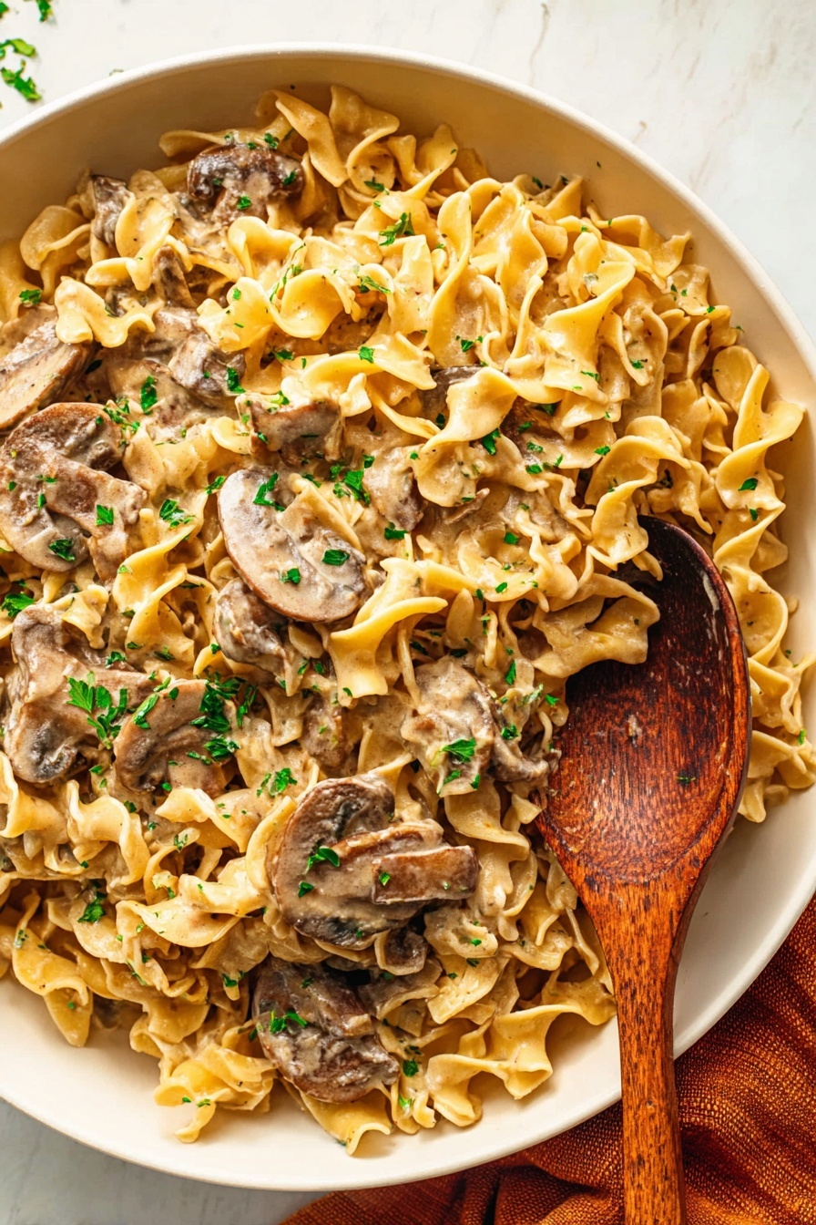 A large white bowl filled with several layers of wavy egg noodles mixed with thick slices of brown mushrooms, all coated in a creamy light beige sauce. Bright green chopped herbs are sprinkled evenly on top, adding a pop of color. A wooden spoon with a rich dark wood grain is placed in the bowl, resting near the right side, lifting some noodles and mushrooms. The bowl sits on a white marbled surface with a folded burnt orange cloth visible at the bottom right corner. Photo taken with an iphone --ar 2:3 --v 7