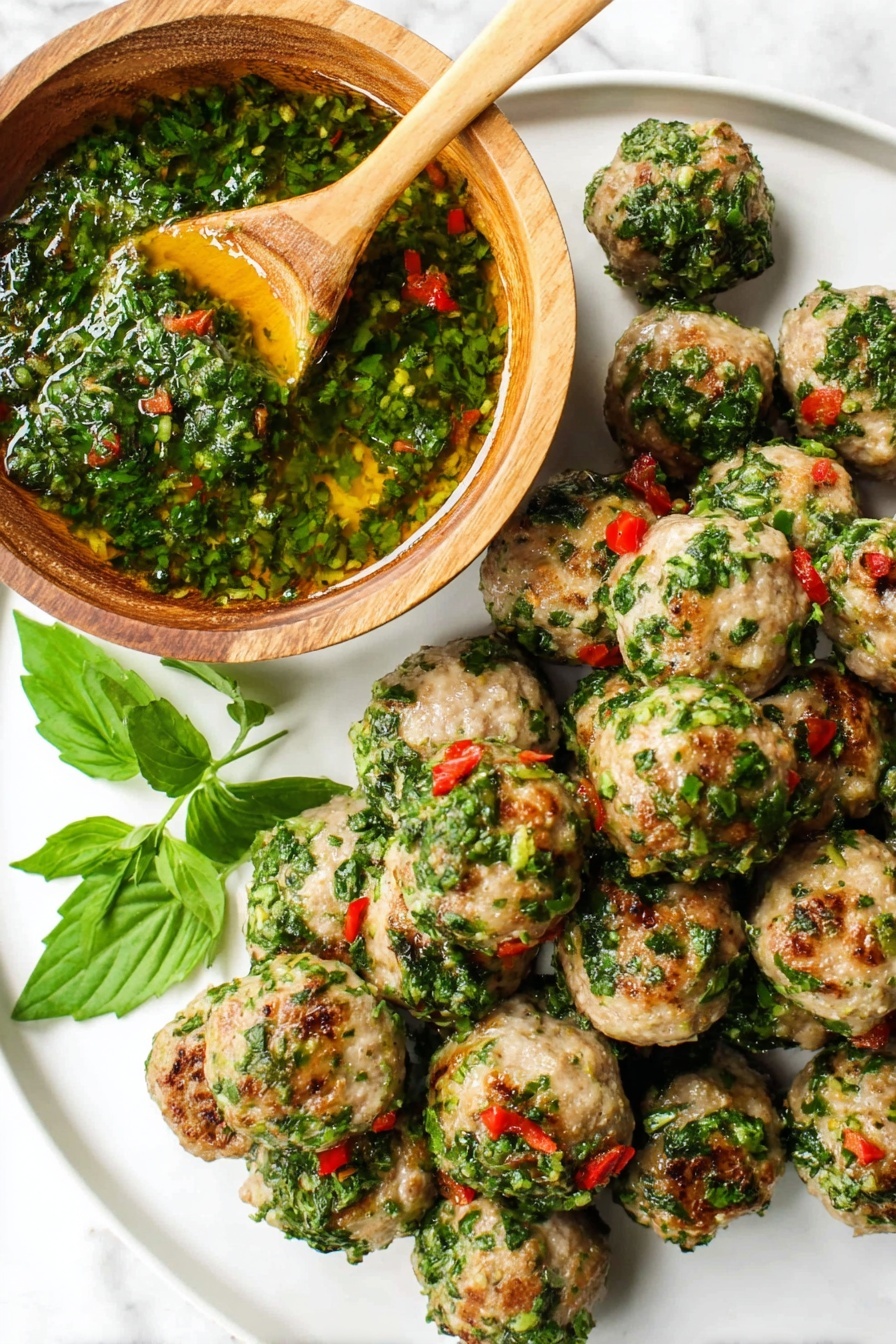 The image shows a white plate filled with small, round meatballs that have a light brown cooked color and are topped with vibrant green chopped herbs mixed with small pieces of red chili, giving a fresh and spicy look. On the top left of the plate, there is a wooden bowl filled with a thick, green herb sauce, glistening with oil, with a wooden spoon resting inside it. Some green herb leaves are scattered around the plate, adding more color contrast. The whole scene is set on a white marbled surface. photo taken with an iphone --ar 2:3 --v 7