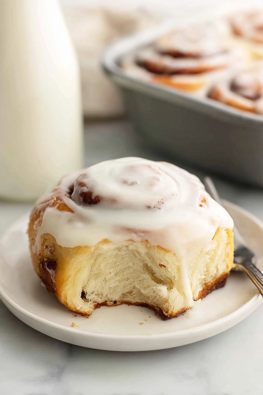 A single cinnamon roll sits on a small white plate placed on a white marbled surface. The roll has two visible layers: a golden-brown baked dough base with some darker cinnamon swirls inside, and a thick, creamy white icing layer spread on top, slightly dripping down the sides. The roll shows a soft, fluffy texture where a small bite has been taken out. In the background, part of a white milk bottle and a gray baking pan holding more cinnamon rolls are visible, all on the same white marbled surface. Photo taken with an iphone --ar 2:3 --v 7