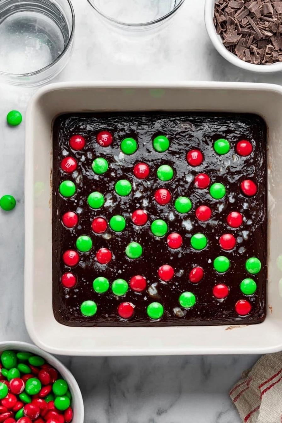 The image shows a grid of square dessert bars laid close together on a white marbled surface. Each bar has two clear layers: a dark brown, dense base that looks soft and moist, and a thick white layer on top that is covered with bright red and green round candies, small chocolate pieces, and red, green, and white sprinkles that add a festive look. The top layer is colorful and textured, with the candies scattered unevenly but filling each square well. Some of the candies are broken or partially embedded, adding more texture to the surface. photo taken with an iphone --ar 2:3 --v 7