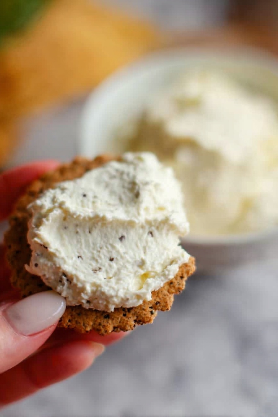 A white bowl filled with a thick, creamy white spread swirled in a circle, with a drizzle of yellow oil on top creating a spiral pattern. On one side of the bowl, there are fresh green basil leaves gathered together. Around the bowl, there are almond slivers scattered on the top right, a squeezed lemon half on the top left, and some fresh basil leaves on the bottom left, all placed on a white marbled surface. Photo taken with an iphone --ar 2:3 --v 7